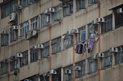 A tall, beige apartment building with multiple identical windows, each equipped with air conditioning units. Clothes are hung to dry on clotheslines outside several windows, adding a sense of daily life to the scene.
