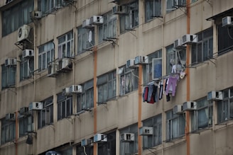 A tall, beige apartment building with multiple identical windows, each equipped with air conditioning units. Clothes are hung to dry on clotheslines outside several windows, adding a sense of daily life to the scene.