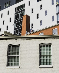 The image depicts a section of an urban building facade with large windows and a brick chimney. The lower part features a white brick wall with two windows having shutters. In the background, there's a taller modern building with contrasting black and white sections and multiple windows.