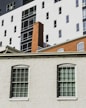 The image depicts a section of an urban building facade with large windows and a brick chimney. The lower part features a white brick wall with two windows having shutters. In the background, there's a taller modern building with contrasting black and white sections and multiple windows.