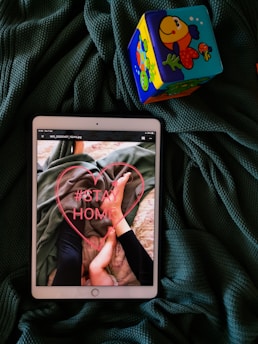 An iPad screen displays an image of a person holding a child's feet. The words '#STAY HOME' are written in red inside a heart. The iPad is placed on a dark green textured fabric, next to a colorful toy block featuring fish illustrations.