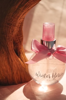 A transparent round bottle labeled 'Body Mist Winter Berries' is adorned with a shiny pink ribbon. The bottle has a pink spray cap and stands against a wooden textured background, casting a gentle shadow.