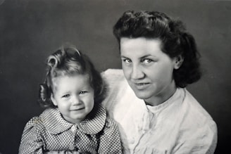 A black and white photograph of a woman and a child posing closely together. The woman has short wavy hair and is looking at the camera with a slight smile. The child, who has curly hair styled in curls, is also looking at the camera and wearing a checkered dress with a collar.
