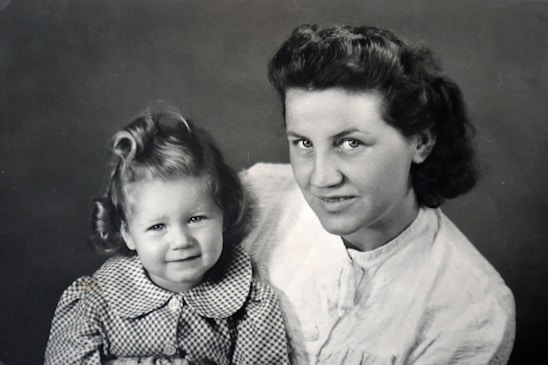 A black and white photograph of a woman and a child posing closely together. The woman has short wavy hair and is looking at the camera with a slight smile. The child, who has curly hair styled in curls, is also looking at the camera and wearing a checkered dress with a collar.