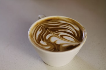 A cup of coffee with intricate latte art on the surface, creating a swirling pattern of brown and white. The cup is placed on a light-colored countertop.