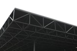 A large, industrial metal structure extending into the sky. The geometric framework consists of a series of triangular sections and beams, creating an intricate pattern. The background is overcast, with a predominantly grey sky that contrasts with the dark metal of the structure.