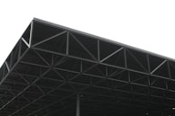 A large, industrial metal structure extending into the sky. The geometric framework consists of a series of triangular sections and beams, creating an intricate pattern. The background is overcast, with a predominantly grey sky that contrasts with the dark metal of the structure.