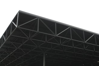 A large, industrial metal structure extending into the sky. The geometric framework consists of a series of triangular sections and beams, creating an intricate pattern. The background is overcast, with a predominantly grey sky that contrasts with the dark metal of the structure.