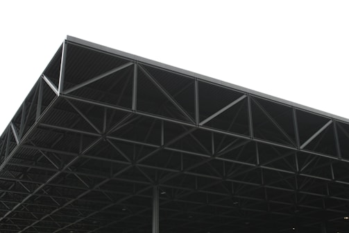 A large, industrial metal structure extending into the sky. The geometric framework consists of a series of triangular sections and beams, creating an intricate pattern. The background is overcast, with a predominantly grey sky that contrasts with the dark metal of the structure.