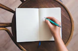 A person's hand is holding a turquoise pen poised over an open lined notebook. The notebook rests on a round wooden chair, suggesting a casual setting. The hand is ready to write and the notebook pages are blank.