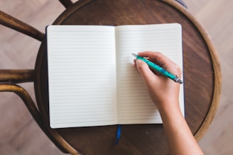 A person's hand is holding a turquoise pen poised over an open lined notebook. The notebook rests on a round wooden chair, suggesting a casual setting. The hand is ready to write and the notebook pages are blank.