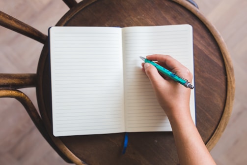 A person's hand is holding a turquoise pen poised over an open lined notebook. The notebook rests on a round wooden chair, suggesting a casual setting. The hand is ready to write and the notebook pages are blank.