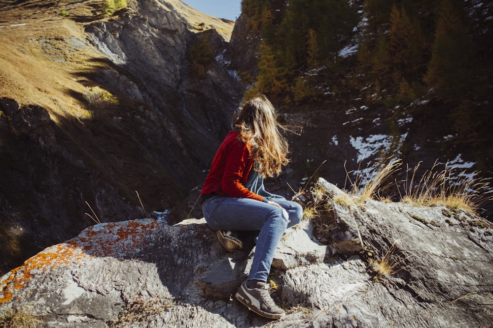 woman in red jacket and blue denim jeans sitting on rock near river during daytime