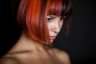 A person with a short bob hairstyle featuring vibrant red and orange hues. The lighting is low, focusing on the rich colors of the hair and the contemplative expression on the person's face. The background is dark, enhancing the contrast.