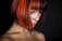 A person with a short bob hairstyle featuring vibrant red and orange hues. The lighting is low, focusing on the rich colors of the hair and the contemplative expression on the person's face. The background is dark, enhancing the contrast.