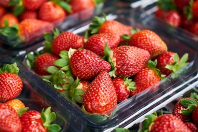 Bright red strawberries are neatly packed in clear plastic containers. The strawberries are fresh, with green leaves still attached, and are arranged with multiple containers visible in the background.