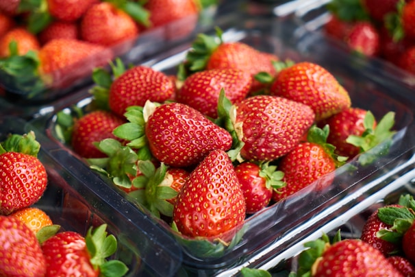 Bright red strawberries are neatly packed in clear plastic containers. The strawberries are fresh, with green leaves still attached, and are arranged with multiple containers visible in the background.