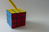 A Rubik's cube with blue, red, and yellow faces sits on a plain surface. A yellow pen is pointed towards the center of the yellow face.