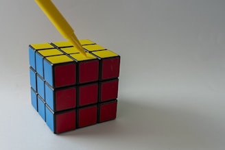 A Rubik's cube with blue, red, and yellow faces sits on a plain surface. A yellow pen is pointed towards the center of the yellow face.