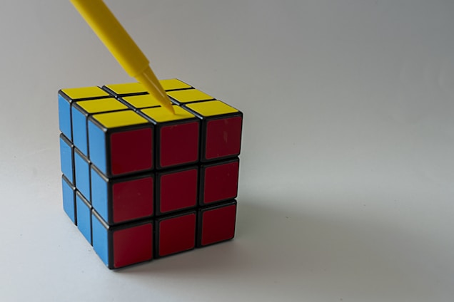 A Rubik's cube with blue, red, and yellow faces sits on a plain surface. A yellow pen is pointed towards the center of the yellow face.