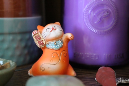 An orange Maneki-neko cat figurine with a raised paw and wearing a beaded blue necklace is placed among a collection of items including a purple container and other small objects.