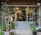 A quaint storefront with large glass doors surrounded by various potted plants, including leafy greens and colorful flowers. Inside, more plants are visible, creating a lush and inviting atmosphere. A chalkboard with writing leans against the wall to the left, adding a cozy, rustic feel.