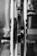 A close-up view of a bike trainer setup, showing the rear wheel mounted on the trainer with a focus on the metallic parts. One can see the leg of a person wearing sneakers, suggesting use for cycling or training indoors.