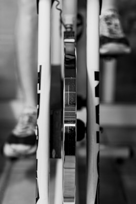 A close-up view of a bike trainer setup, showing the rear wheel mounted on the trainer with a focus on the metallic parts. One can see the leg of a person wearing sneakers, suggesting use for cycling or training indoors.