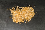 A pile of yellow popcorn kernels scattered on a dark textured surface. The kernels are small, smooth, and glossy, with a mix of yellow and translucent hues.