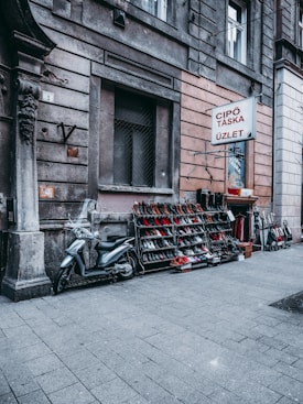 A street scene featuring a store selling shoes with a variety of footwear displayed on racks. A parked scooter is visible next to the racks. The storefront includes a sign with text in a foreign language and is set against an urban backdrop with textured walls.