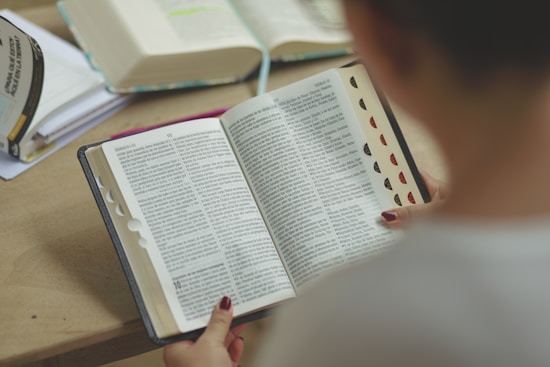 A person is sitting at a table reading a thick, open book. The pages are filled with text, and there are tabs sticking out for easy navigation. Two other open books are visible in the background, along with a notebook.