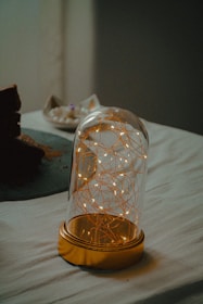 A glass dome encases delicate strands of fairy lights, emitting a soft, warm glow. The dome is set on a table, which is covered with a light-colored cloth. To the right, some white and brown items appear out of focus, possibly decorative or edible elements.