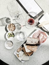 A charcuterie board is laid out with an assortment of crackers, cheeses, nuts, and cold cuts arranged neatly on a platter. Two wine glasses, one empty and one with red wine, are placed on a speckled white table. A menu is partially visible in the upper part of the image next to a wooden fork on a napkin. A small bowl contains almonds, and another bowl has green olives.