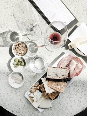 A charcuterie board is laid out with an assortment of crackers, cheeses, nuts, and cold cuts arranged neatly on a platter. Two wine glasses, one empty and one with red wine, are placed on a speckled white table. A menu is partially visible in the upper part of the image next to a wooden fork on a napkin. A small bowl contains almonds, and another bowl has green olives.