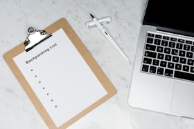 A clipboard with a paper titled 'Backpacking List' lies on a white marble surface. Beside it, there is a white pen and a laptop with a visible keyboard. The scene suggests organization or planning, possibly for a trip.