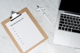 A clipboard with a paper titled 'Backpacking List' lies on a white marble surface. Beside it, there is a white pen and a laptop with a visible keyboard. The scene suggests organization or planning, possibly for a trip.