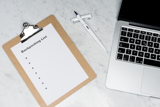 A clipboard with a paper titled 'Backpacking List' lies on a white marble surface. Beside it, there is a white pen and a laptop with a visible keyboard. The scene suggests organization or planning, possibly for a trip.