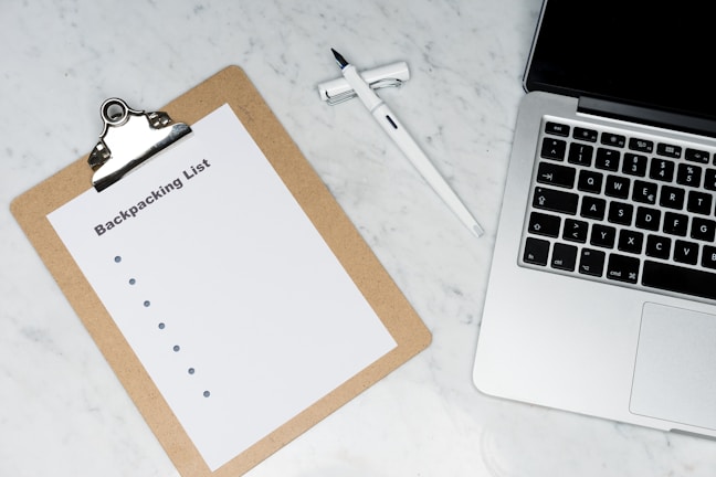 A clipboard with a paper titled 'Backpacking List' lies on a white marble surface. Beside it, there is a white pen and a laptop with a visible keyboard. The scene suggests organization or planning, possibly for a trip.