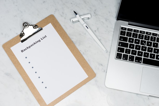 A clipboard with a paper titled 'Backpacking List' lies on a white marble surface. Beside it, there is a white pen and a laptop with a visible keyboard. The scene suggests organization or planning, possibly for a trip.