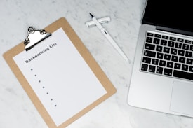 A clipboard with a paper titled 'Backpacking List' lies on a white marble surface. Beside it, there is a white pen and a laptop with a visible keyboard. The scene suggests organization or planning, possibly for a trip.