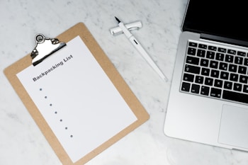 A clipboard with a paper titled 'Backpacking List' lies on a white marble surface. Beside it, there is a white pen and a laptop with a visible keyboard. The scene suggests organization or planning, possibly for a trip.