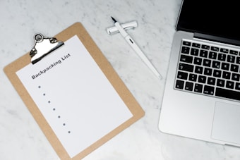 A clipboard with a paper titled 'Backpacking List' lies on a white marble surface. Beside it, there is a white pen and a laptop with a visible keyboard. The scene suggests organization or planning, possibly for a trip.