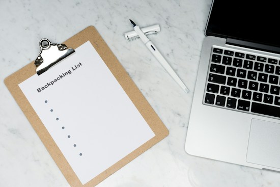 A clipboard with a paper titled 'Backpacking List' lies on a white marble surface. Beside it, there is a white pen and a laptop with a visible keyboard. The scene suggests organization or planning, possibly for a trip.