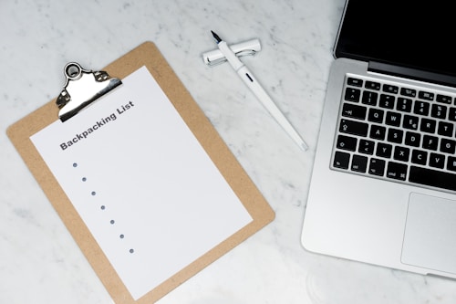 A clipboard with a paper titled 'Backpacking List' lies on a white marble surface. Beside it, there is a white pen and a laptop with a visible keyboard. The scene suggests organization or planning, possibly for a trip.