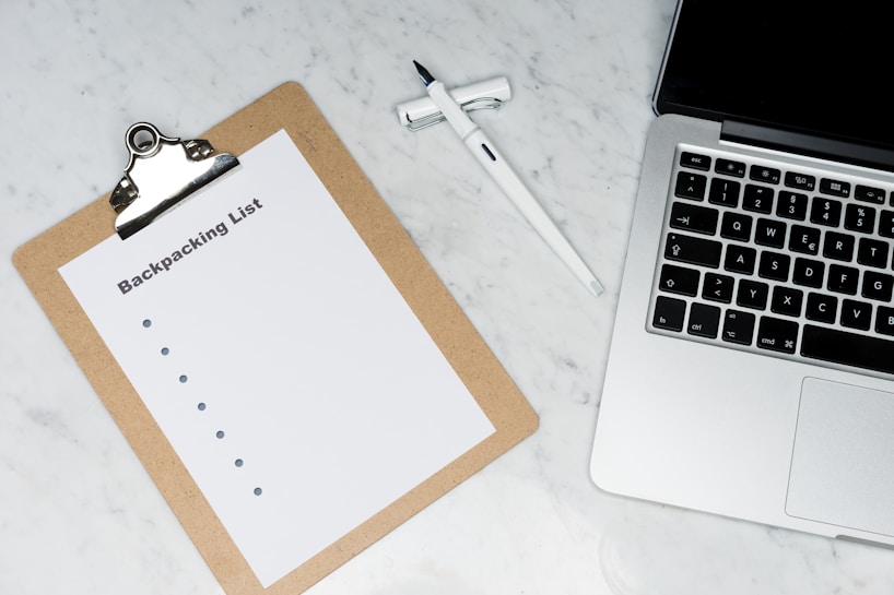 A clipboard with a paper titled 'Backpacking List' lies on a white marble surface. Beside it, there is a white pen and a laptop with a visible keyboard. The scene suggests organization or planning, possibly for a trip.