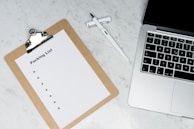 A blank packing list on a clipboard is placed on a marble surface beside a pen and an open laptop.