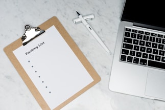 A blank packing list on a clipboard is placed on a marble surface beside a pen and an open laptop.