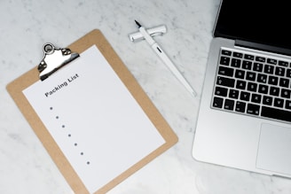 A blank packing list on a clipboard is placed on a marble surface beside a pen and an open laptop.