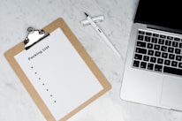 A blank packing list on a clipboard is placed on a marble surface beside a pen and an open laptop.