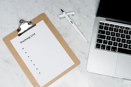 Close-up of a hand checking off items on a printed packing checklist beside a laptop